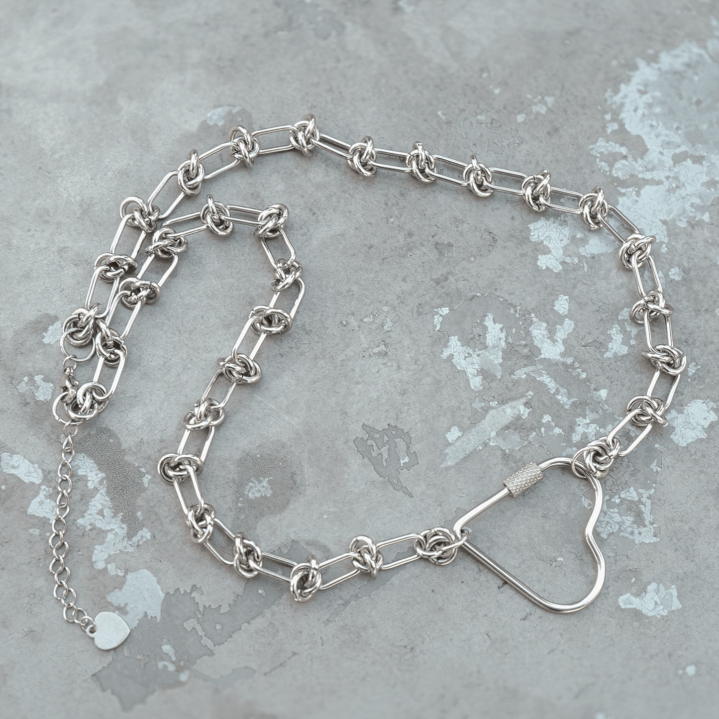 soft but deadly collection stainless necklace featuring silver knotted chain links and silver heart carabiner pendant on a concreate background