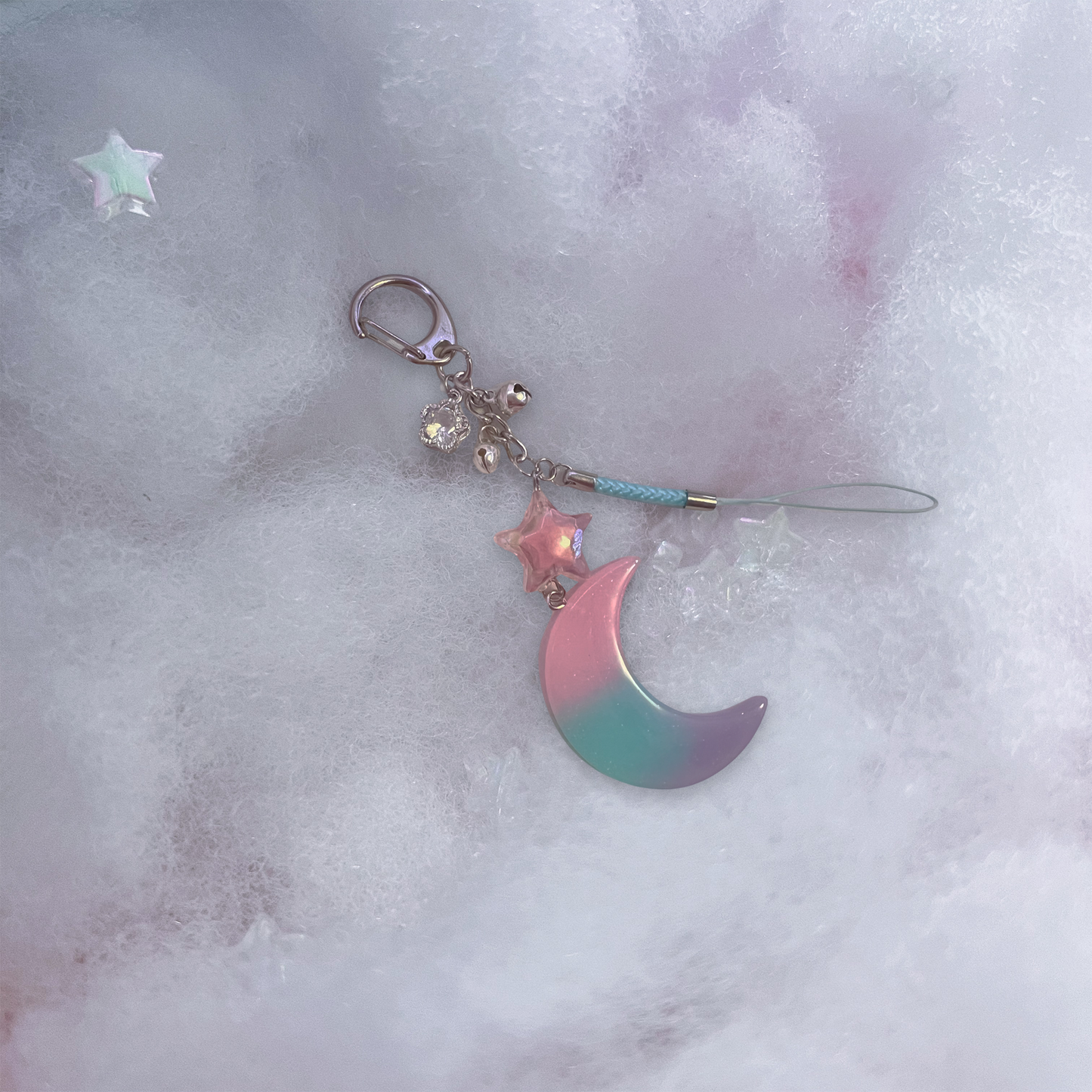 Specialty keychain featuring a sparkly resin moon charm with pastel colors and connecting star charm, with small bells and crystal flower charm and braided phone strap.