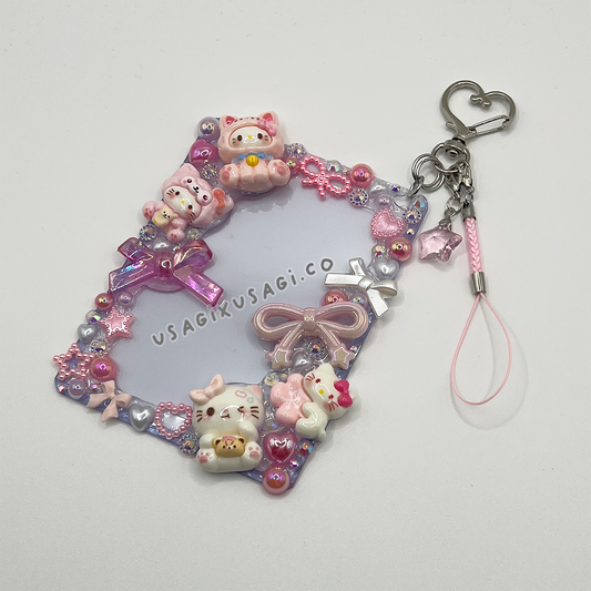A decorative heart-shaped keychain case with pink and lavender glitter, small 3D Hello Kitty figures, bows, rhinestones, and a silver clasp with a pink loop strap, photographed on a plain light background.